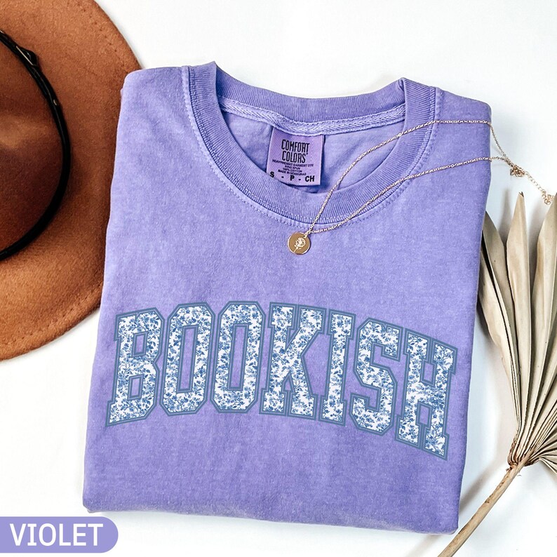 May include: A violet-colored t-shirt with the word "BOOKISH" in a floral design across the front. A gold necklace with a pendant is draped over the shirt. A brown hat and a dried palm leaf are in the background.