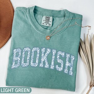 May include: A light green t-shirt with the word "BOOKISH" in large, floral-patterned letters. A delicate silver necklace with a pendant rests on the shirt. The shirt is labeled "LIGHT GREEN" at the bottom.