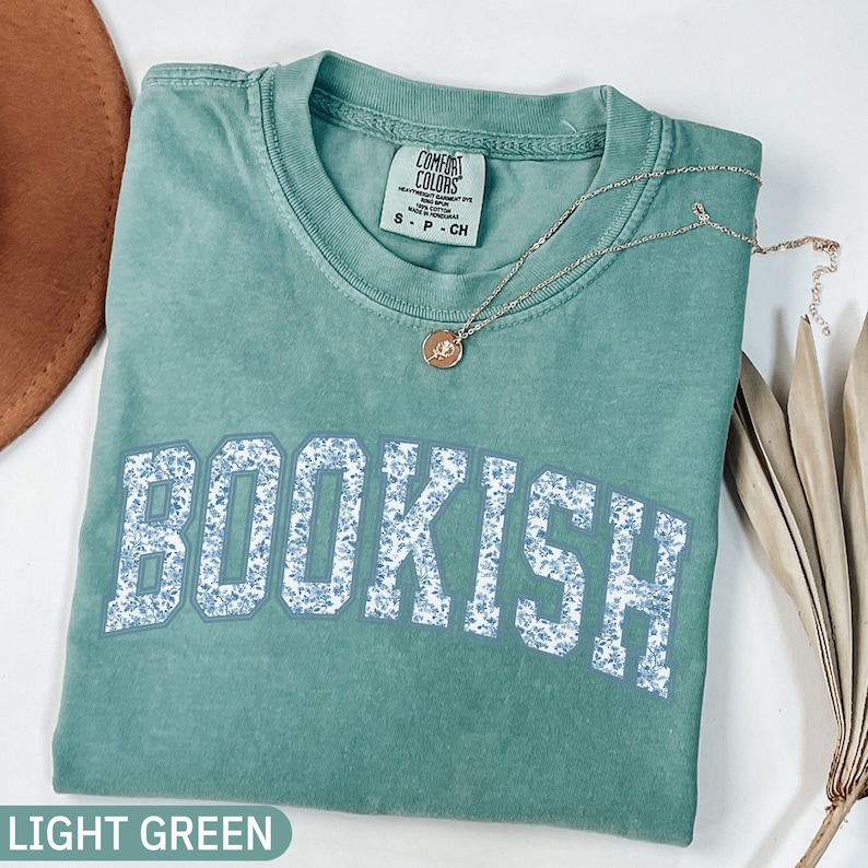 May include: A light green t-shirt with the word "BOOKISH" in large, floral-patterned letters. A delicate silver necklace with a pendant rests on the shirt. The shirt is labeled "LIGHT GREEN" at the bottom.
