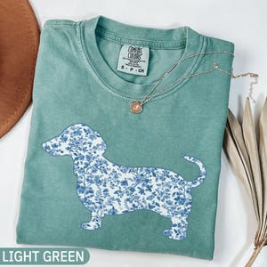 May include: Light green Comfort Colors t-shirt featuring a dachshund silhouette filled with a blue floral pattern. A delicate silver necklace with a pendant rests on the shirt. The text "LIGHT GREEN" is at the bottom.