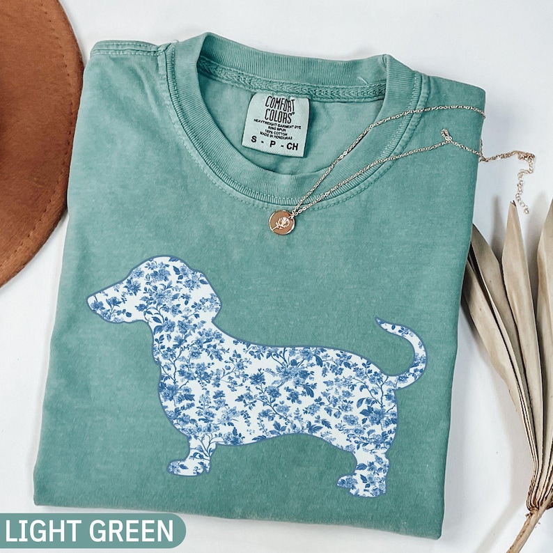 May include: Light green Comfort Colors t-shirt featuring a dachshund silhouette filled with a blue floral pattern. A delicate silver necklace with a pendant rests on the shirt. The text "LIGHT GREEN" is at the bottom.