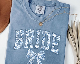 May include: A light blue t-shirt with the word "BRIDE" in white letters outlined in blue. The letters are covered in a blue floral pattern. A white bow with a blue floral pattern is below the word "BRIDE". The shirt has a tag that says "COMFORT COLORS" and "S - P - CH".