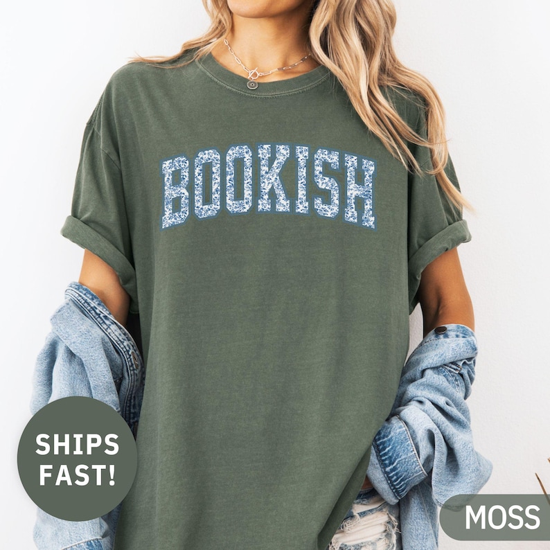 May include: A moss green t-shirt with the word "BOOKISH" in a blue floral design across the chest. The shirt has short sleeves and a relaxed fit. A denim jacket is draped over the shoulders.
