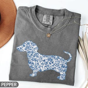 May include: A gray Comfort Colors t-shirt featuring a blue floral dachshund design. The shirt has a crew neck and short sleeves. A gold necklace with a pendant is draped on the shirt. The word "PEPPER" is at the bottom left.