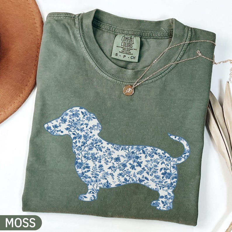 May include: A moss green t-shirt featuring a blue floral dachshund design. The shirt has a crew neck and short sleeves. A gold necklace with a pendant is draped on the shirt. The word "MOSS" is printed in a white rectangle at the bottom.