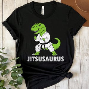 May include: Black t-shirt featuring a green dinosaur in a white karate uniform with a black belt. The dinosaur is in a fighting stance. The word "JITSUSAURUS" is printed in white below the dinosaur.