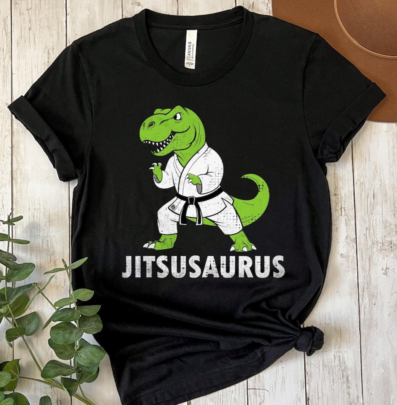 May include: Black t-shirt featuring a green dinosaur in a white karate uniform with a black belt. The dinosaur is in a fighting stance. The word "JITSUSAURUS" is printed in white below the dinosaur.