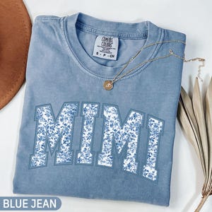 Könnte beinhalten: Ein blaues Jeans-T-Shirt mit einem weißen Blumenmuster, auf dem in einer College-Schriftart "MIMI" steht. Das Shirt ist aus einem weichen, bequemen Stoff und perfekt für den Alltag.