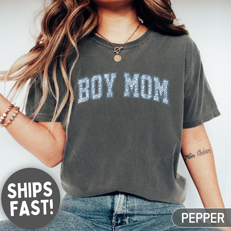 K&ouml;nnte beinhalten: Dunkelgraues T-Shirt mit dem Text "BOY MOM" in hellblauer, floral gemusterter Schrift. Das Shirt hat einen Rundhalsausschnitt und scheint aus einem weichen, bequemen Material zu sein. Das Wort "PEPPER" steht unten rechts im Bild.