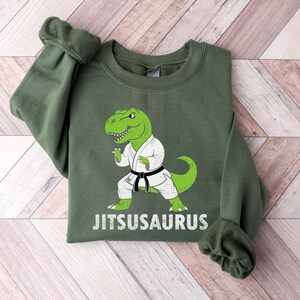 May include: Green sweatshirt featuring a cartoon green dinosaur in a karate uniform with a black belt. The text "Jitsusaurus" is printed in white below the dinosaur. The sweatshirt has a crew neck and long sleeves.