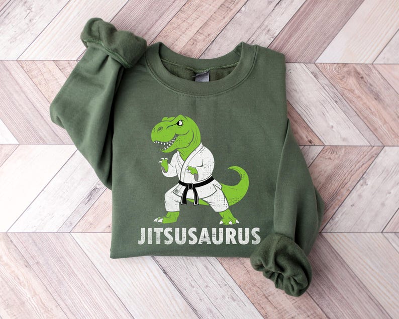 May include: Green sweatshirt featuring a cartoon green dinosaur in a karate uniform with a black belt. The text "Jitsusaurus" is printed in white below the dinosaur. The sweatshirt has a crew neck and long sleeves.