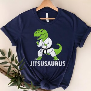 May include: Navy blue t-shirt featuring a green dinosaur in a karate uniform with a black belt. The dinosaur is in a fighting stance. The text "Jitsusaurus" is printed in white below the dinosaur.