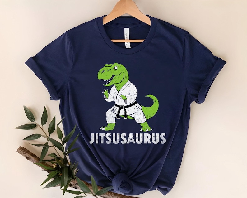 May include: Navy blue t-shirt featuring a green dinosaur in a karate uniform with a black belt. The dinosaur is in a fighting stance. The text "Jitsusaurus" is printed in white below the dinosaur.