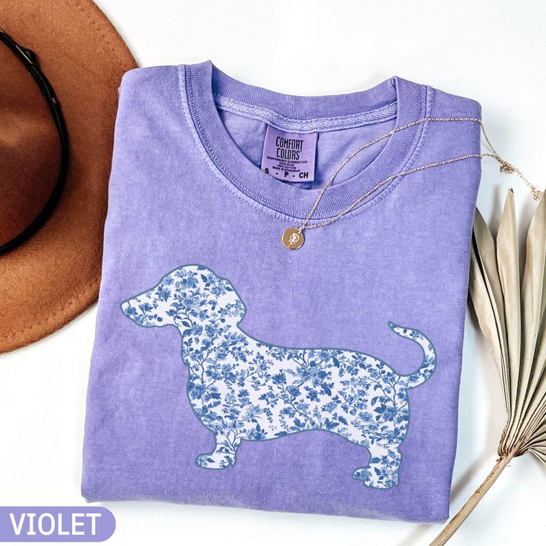 May include: A violet Comfort Colors t-shirt featuring a floral dachshund design in shades of blue. The shirt has a crew neck and short sleeves. A gold necklace with a small pendant is draped across the shirt. The word "VIOLET" is printed at the bottom.