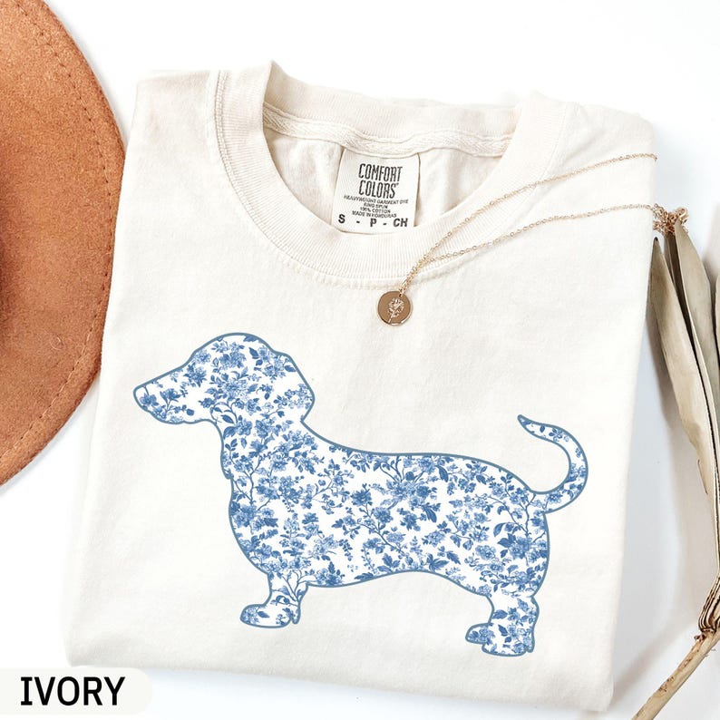 May include: Ivory-colored t-shirt featuring a blue floral dachshund design. The dog silhouette is filled with a delicate blue floral pattern. The shirt has a crew neck and short sleeves. A gold necklace with a pendant is also visible.