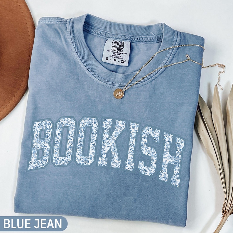 May include: A blue jean colored t-shirt with the word "BOOKISH" in white letters with a floral pattern. A gold necklace with a pendant is draped across the shirt. The shirt has a Comfort Colors tag.
