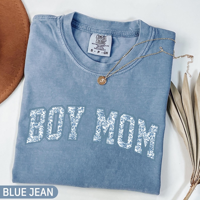 K&ouml;nnte beinhalten: Ein blaues Comfort Colors T-Shirt mit der Aufschrift "BOY MOM" in wei&szlig;en Blockbuchstaben mit floralem Muster. Eine goldene Halskette mit Anh&auml;nger liegt &uuml;ber dem Shirt. Das Shirt ist gefaltet und auf einer wei&szlig;en Oberfl&auml;che platziert.