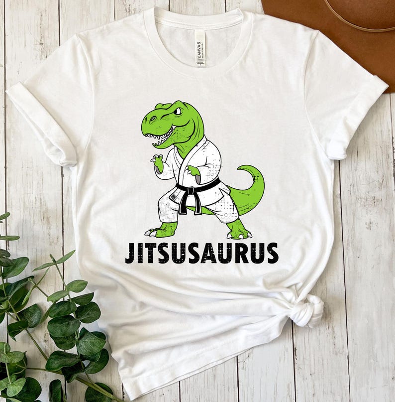 May include: White t-shirt featuring a green dinosaur in a karate uniform with a black belt. The text "JITSUSAURUS" is printed in black below the dinosaur illustration. The shirt is made of a soft, comfortable material.