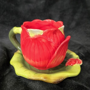 May include: A ceramic cup and saucer set shaped like a red tulip flower. The cup features a bright red petal exterior with a green stem-shaped handle. The saucer is designed as a green leaf with a small red bud accent on the side.