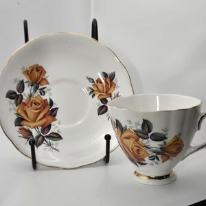 May include: White teacup and saucer set with a gold rim and a floral rose design in shades of yellow, orange, and green. The teacup has a curved handle and sits on a small foot. The saucer is round.