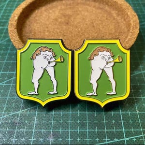 Könnte beinhalten: Zwei Emaille-Anstecknadeln mit einem skurrilen Design einer Figur, die ein Horn spielt. Die Anstecknadeln haben einen grünen Hintergrund mit gelbem Rand und eine Figur mit braunen Haaren. Die Anstecknadeln liegen auf einer grünen Rasteroberfläche.