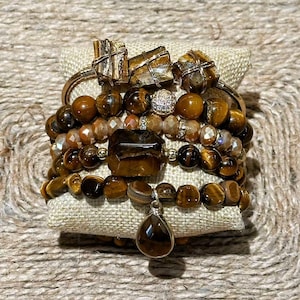 Puede incluir: Una colección de pulseras y anillos apilados con cuentas de ojo de tigre y detalles dorados. Las pulseras incluyen varios tamaños y formas de cuentas, con un colgante en forma de lágrima. Los anillos tienen diseños geométricos.