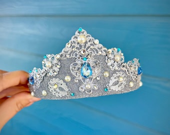 May include: A silver and gray crown with intricate lace detailing, adorned with faux pearls and blue gemstones. The crown has a glittery finish and is set against a blue background.