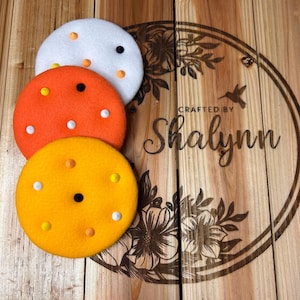 May include: Three colorful, round coasters in white, orange, and yellow, each adorned with small, contrasting-colored dots. The coasters are arranged on a wooden surface with the text "Crafted by Shalynn" within a floral design.