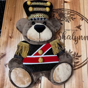 May include: A plush brown teddy bear dressed as a toy soldier, wearing a black hat with gold trim and buttons, a black jacket with gold fringe epaulets, and a red and white sash with two gold buttons across the chest, featuring a soft snout.