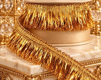May include: A spool of metallic gold fringe trim featuring a dense, layered design of thin, reflective strands attached to a woven header. The trim is wrapped around a circular cardboard core, showcasing a bright, shiny finish throughout.