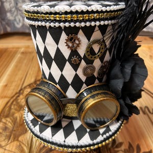 May include: A steampunk-style top hat with a black and white diamond pattern. The hat features decorative gears, gold-rimmed goggles, black feathers, and a pearl and gold trim. The base of the hat is also decorated with a diamond pattern.