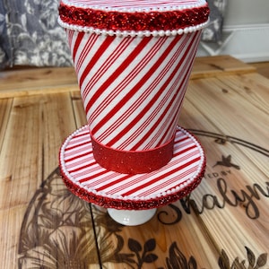 May include: A decorative candy cane-striped top hat with a matching base. The hat and base are adorned with red glitter ribbon and white pearl-like beads. The hat sits on a small white pedestal.