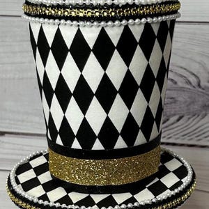 May include: A decorative top hat with a black and white diamond pattern. The hat features a gold glitter band and is trimmed with pearls and gold beads. The brim also has a black and white diamond pattern.
