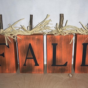 May include: Four rectangular orange wooden blocks arranged in a row, each featuring a single black letter spelling the word FALL. Each block is topped with a brown twig stem and light tan raffia fibers, creating a textured, rustic appearance.