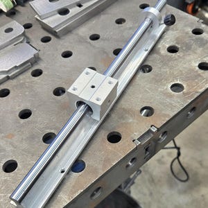 May include: A silver linear bearing rail with a silver linear bearing block attached to it. The rail is mounted on a metal workbench.