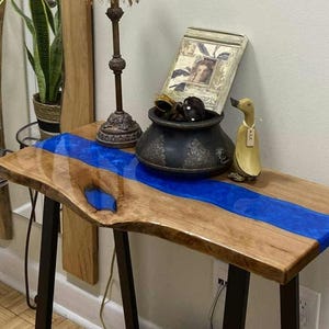 Può includere: Tavolo consolle in legno con bordo naturale e fiume in resina blu. Il tavolo, lungo circa 100 cm, ha gambe in metallo nero ed è decorato con una lampada, una cornice e oggetti decorativi.