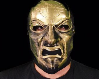 May include: A gold and green mask with a detailed design and a menacing expression. The mask has a symbol on the forehead and a large mouth with sharp teeth.