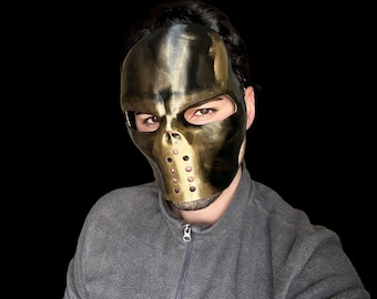 May include: A gold-colored skull mask with a metal grill over the mouth and eye holes. The mask is worn by a person with a gray sweater.