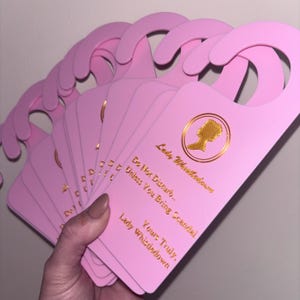 May include: A collection of pink door hangers with a curved top. Each hanger features gold text and a gold silhouette of a woman's profile. The text reads "Do Not Disturb... Unless You Bring Scandal. Yours Truly, Lady Whistledown."