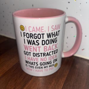 May include: A white ceramic mug with a pink interior and handle. The exterior features black and pink text that reads: I CAME, I SAW I FORGOT WHAT I WAS DOING WENT BACK GOT DISTRACTED I HAVE NO IDEA WHATS GOING ON IS THIS EVEN MY MUG NOW I HAVE TO PEE.