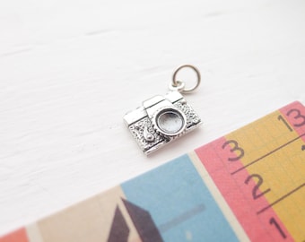 Può includere: Un piccolo ciondolo a forma di macchina fotografica color argento con un design dettagliato, tra cui un obiettivo ed elementi decorativi. Il ciondolo ha un anello per attaccarlo a una collana o un braccialetto. Sullo sfondo, un righello colorato con sezioni blu, rosa e gialle.