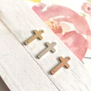 May include: Three metal cross charms in gold, silver, and rose gold. The charms are flat and have a simple design.