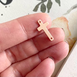 May include: A small, rose gold-colored metal cross charm with two holes for attaching to jewelry. The cross is flat and has a slightly textured surface.