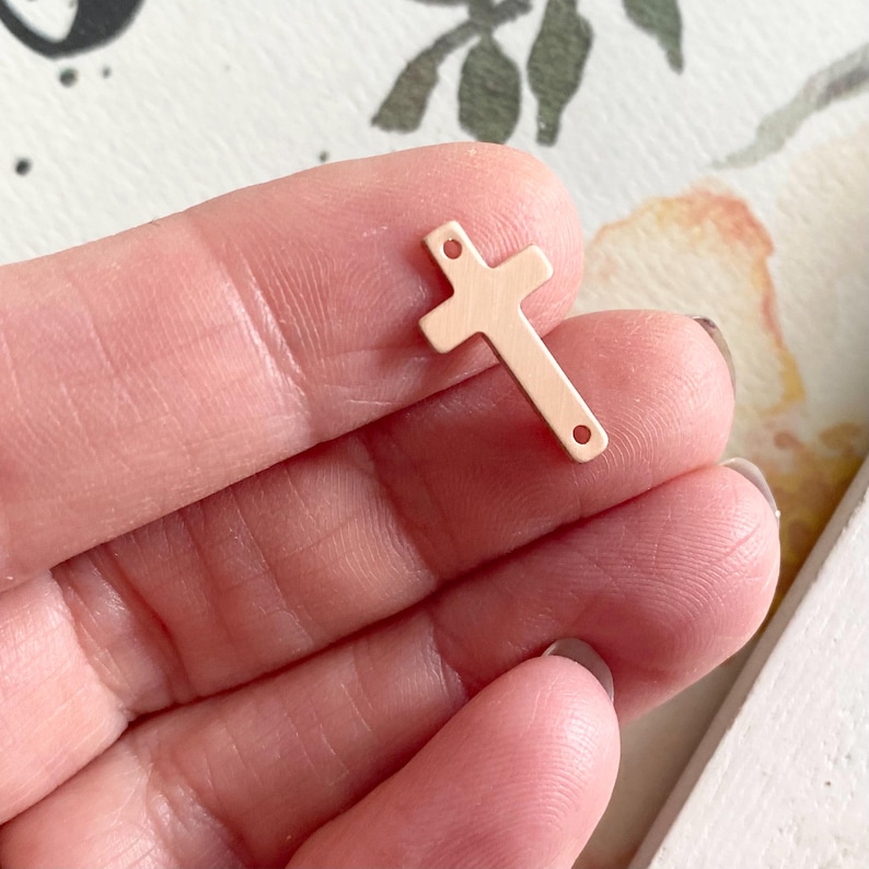 May include: A small, rose gold-colored metal cross charm with two holes for attaching to jewelry. The cross is flat and has a slightly textured surface.