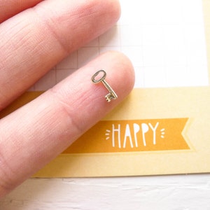 May include: A small, silver-colored key charm with a simple design. The key is resting on a finger, and the background is a yellow surface with the word "HAPPY" written in white.
