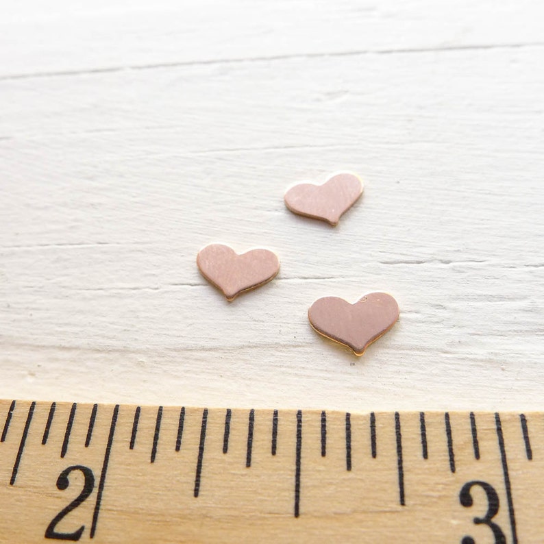 May include: Three small pink heart-shaped metal charms on a white wooden surface with a ruler in the foreground.