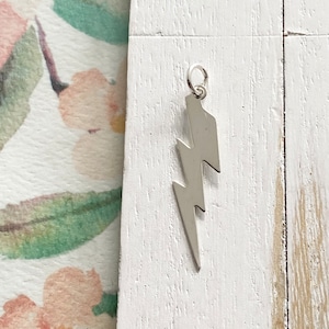 May include: Silver lightning bolt charm pendant. The charm is a simple, flat design with a smooth finish.