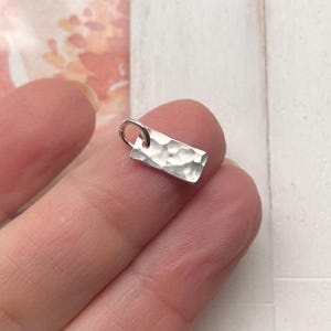 May include: A small, silver, rectangular charm with a hammered texture. The charm has a jump ring for attaching to a necklace or bracelet.