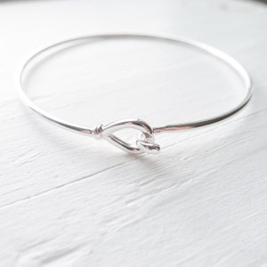 May include: A silver bangle bracelet with a knot design. The bracelet is open and has a simple, elegant look.