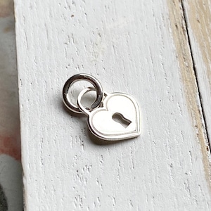 May include: A silver heart-shaped charm with a keyhole cutout. The charm has a ring attached to the top for attaching to a necklace or bracelet.